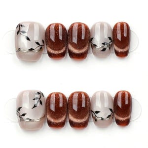 May include: Set of artificial nails in two rows. Each nail is oval-shaped with a glossy finish. Some nails are a light beige with black leaf designs and silver accents. Others are a deep brown with a shimmering effect.