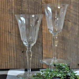 May include: Two clear, stemmed wine glasses with etched floral designs. The glasses have a flared bowl shape and sit on a round base. The background is a rustic wooden surface.