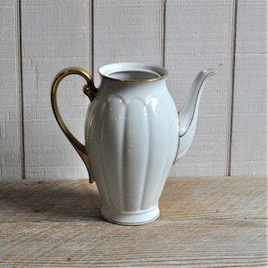 May include: White porcelain coffee pot with a gold handle and trim. The pot has a fluted design and a curved spout. The handle is a curved, ornate design. The pot is set against a white wooden background.