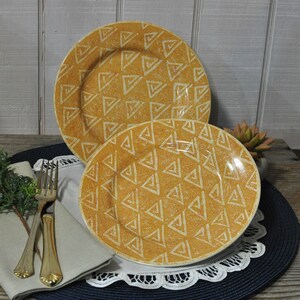 May include: Two yellow ceramic plates with a geometric pattern of triangles. The plates are nested together, with the smaller plate in front. The plates are on a blue and white tablecloth.
