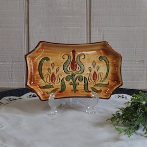 May include: A small, rectangular ceramic tray with a decorative floral design in shades of red, green, and yellow. The tray has a rustic, hand-painted look and is perfect for serving snacks or small items.