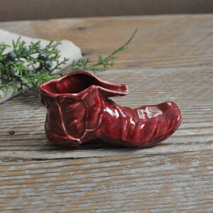 May include: A small, red ceramic boot shaped like a shoe. The boot is glazed and has a textured surface.