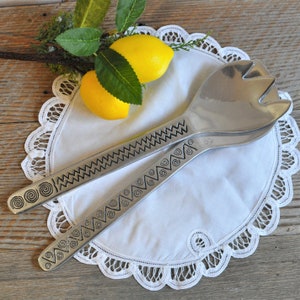 May include: A set of two silver salad servers with decorative patterns on a white lace doily. The servers are made of metal and have a shiny finish. The doily is round and has a scalloped edge.
