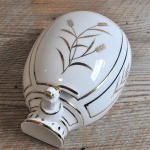 May include: White ceramic bottle stopper with a gold trim and a gold wheat design. The stopper has a spout and a handle.