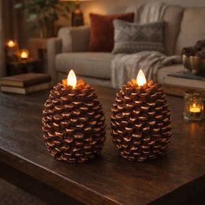 May include: Two decorative pine cone-shaped LED candles with a warm, inviting glow. The candles are bronze-colored and sit on a dark wooden table. The background includes a sofa, lamp, and other home decor items.