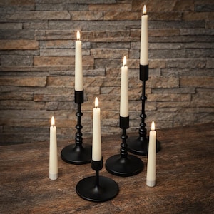 May include: A collection of lit ivory taper candles in black metal candle holders of varying heights. The candle holders have round bases and decorative stems. The candles are placed on a wooden surface against a textured stone wall.