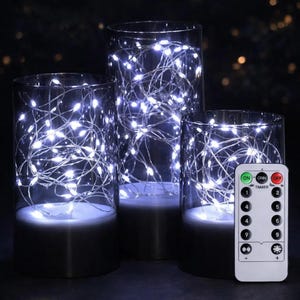 May include: Three clear glass cylinders with white LED string lights inside. The cylinders have a black base and are different heights. A remote control with buttons is in the foreground. The background is dark.