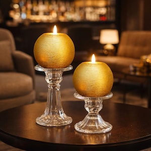 Op de afbeelding: Twee goudkleurige, bolvormige kaarsen met een getextureerd oppervlak, elk op een heldere, sierlijke glazen kandelaar. De kaarsen zijn aangestoken en geven een warme gloed af. De setting is een zwak verlichte kamer met een donkerbruine tafel en een vage achtergrond.