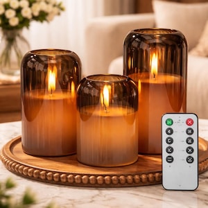 以下が含まれることがあります： さまざまなサイズのガラスホルダーに入った、3つの点灯した琥珀色のピラーキャンドル。 キャンドルは、ビーズの縁が付いた木製のトレイに置かれ、リモコンが横に置かれています。 キャンドルは暖かい光を発しています。