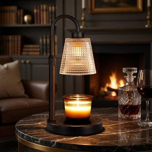 May include: A black candle warmer with a glass shade and a lit candle. The warmer sits on a black marble table, with a decanter and wine glass in the background. The candle is in a glass jar with a gold band.