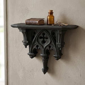 May include: A black wall-mounted shelf featuring intricate architectural detailing with pointed arches and a central cross motif. On the flat top surface sits a small brown leather-bound book, a clear amber glass bottle with a cork, and a set of metal keys.
