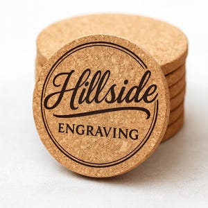 May include: A stack of round cork coasters. The top coaster features the words "Hillside Engraving" in a black, elegant script within a double circular border. The coasters are a natural tan color, with a visible cork texture.