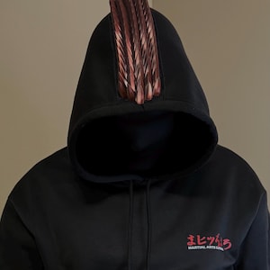 May include: Black hoodie with a decorative, reddish-brown, feather-like embellishment on the hood. The hoodie features a drawstring and a red Japanese text logo that reads "Martial Arts Super".