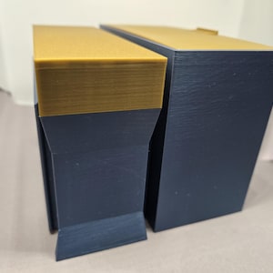 May include: Two geometric, angular objects. The top of each object is a solid gold color, while the sides are a dark navy blue. The objects are positioned next to each other, with one slightly smaller than the other.