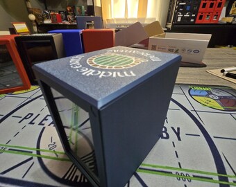 May include: A dark blue card storage box with a clear front panel. The top of the box has a textured surface with the words "Middle Earth" and a circular graphic. Other boxes in various colors are visible in the background.