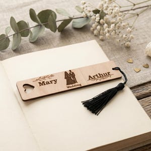 May include: A rectangular light-colored wooden bookmark featuring a laser-engraved design with the names Mary and Arthur, a silhouette of a couple, the word Wedding, and a heart-shaped cutout. A black tassel is attached to the right side.