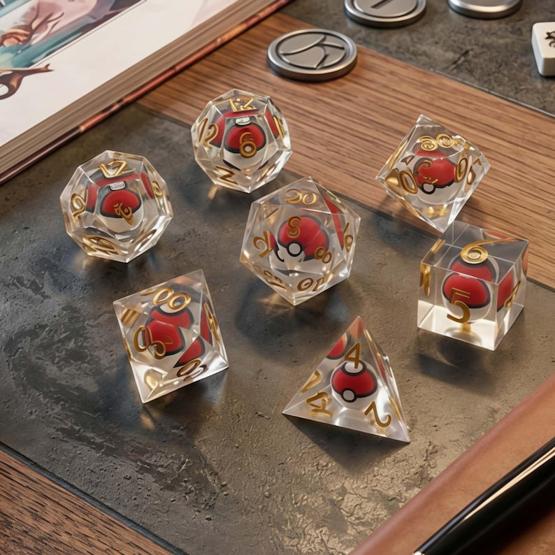 May include: A set of seven polyhedral dice made of clear resin, each containing a miniature red and white spherical object inside. The dice feature gold-colored numbers etched into the surfaces, including d20, d12, d10, d8, d6, and d4 shapes.