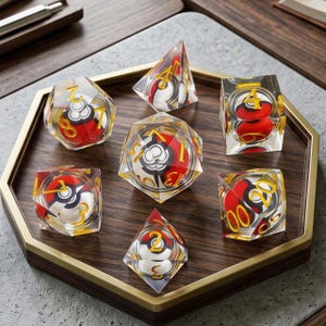 May include: A set of seven clear polyhedral dice, each containing a miniature red, white, and black spherical design inside. The dice feature gold-colored numbers etched into the transparent surfaces, arranged on a wooden hexagonal tray.