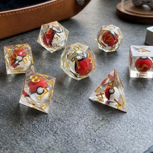 May include: A set of seven clear polyhedral dice, each containing a miniature red and white spherical ball resembling a pokeball. The dice feature gold-colored numbers etched into the transparent resin surfaces, including d20, d12, d10, d8, d6, and d4 shapes.