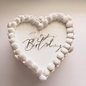 Puede incluir: Un pastel en forma de corazón con glaseado blanco liso y un borde decorativo de remolinos de crema blanca. En el centro, un adorno para pastel de color plateado reflectante muestra el texto Happy Birthday en una fuente cursiva.