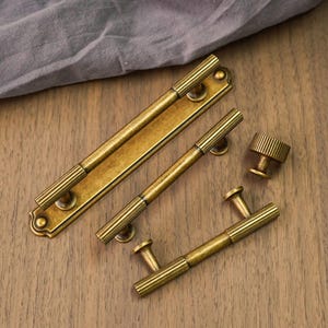 Pode incluir: Uma variedade de ferragens de armário de latão antigo, incluindo puxadores e um botão. Os puxadores têm um design canelado e estão em vários tamanhos. As ferragens estão dispostas em uma superfície de madeira, com um pano cinza ao fundo.