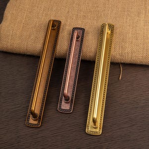 May include: Three metal door pulls in varying shades of bronze and gold. Each pull has a rectangular backplate with a beaded border and a curved handle. The pulls are arranged on a dark wood surface.