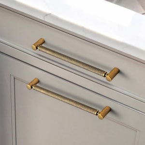 May include: Two brass cabinet pulls with a textured, ridged design. The pulls have a cylindrical bar and cylindrical end caps. The cabinet doors are a light gray color, and the countertop is white with gray veining.