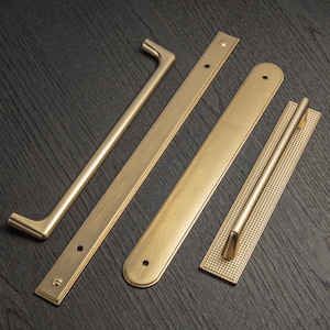 May include: Four brass cabinet hardware pieces are displayed on a dark wood surface. The hardware includes a pull handle, a long bar with screw holes, a bar with rounded ends, and a handle with a dotted pattern.
