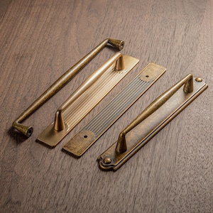 Puede incluir: Cuatro tiradores y manijas de gabinete de latón antiguo en varios diseños. El hardware presenta una variedad de formas, incluyendo tiradores de barra y manijas con placas traseras decorativas. El acabado tiene una apariencia cálida y envejecida.