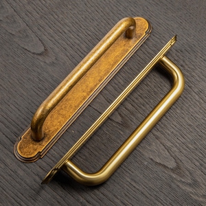 以下が含まれることがあります： 2つのアンティークスタイルのドアハンドル。1つは、湾曲したハンドルが付いた長い長方形のプレートで、もう1つはU字型のハンドルです。どちらもブロンズ仕上げの金属製で、濃い木目の背景に配置されています。