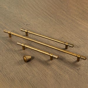 Puede incluir: Un juego de cuatro tiradores de armario de latón antiguo y un pomo a juego. Los tiradores son largos y cilíndricos con detalles acanalados en los extremos. El pomo tiene forma de cono con detalles acanalados similares. El herraje está dispuesto sobre una superficie de madera.