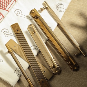 Puede incluir: Una variedad de tiradores y manijas de gabinete de latón antiguo en varios tamaños y diseños. El hardware presenta superficies acanaladas y lisas, con formas en T y rectangulares. Las piezas están dispuestas sobre una tela estampada.