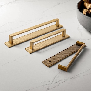 Può includere: Maniglie dorate per mobili con base rettangolare e maniglia a barra. La base presenta un motivo strutturato e puntinato. Due maniglie sono disposte orizzontalmente, e una è smontata.