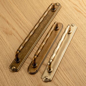 Może przedstawiać: Trzy uchwyty do szafek ze starego brązu z centralnym skręconym wzorem. Uchwyty są w różnych rozmiarach, najdłuższy ma około 30 cm długości. Okucia są umieszczone na jasnej powierzchni z usłojeniem drewna.