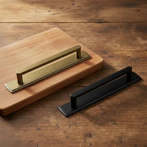 May include: Two rectangular cabinet pulls. One is brass-coloured and mounted on a wooden chopping board. The other is black and rests on a wooden surface. Both have a rectangular base and a rounded handle.