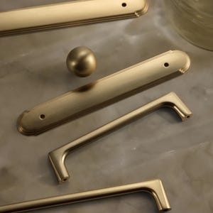 May include: A collection of gold-toned cabinet hardware, including handles, pulls, and a spherical knob. The hardware features a brushed finish and a classic design, suitable for kitchen or bathroom cabinetry. The image showcases various shapes and sizes.
