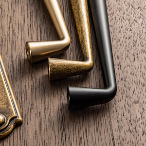 May include: Close-up of three cabinet pulls in different finishes: brass, antique brass, and black. The pulls are L-shaped with a cylindrical grip and are set against a dark wood grain background. The brass pull is a bright gold color.