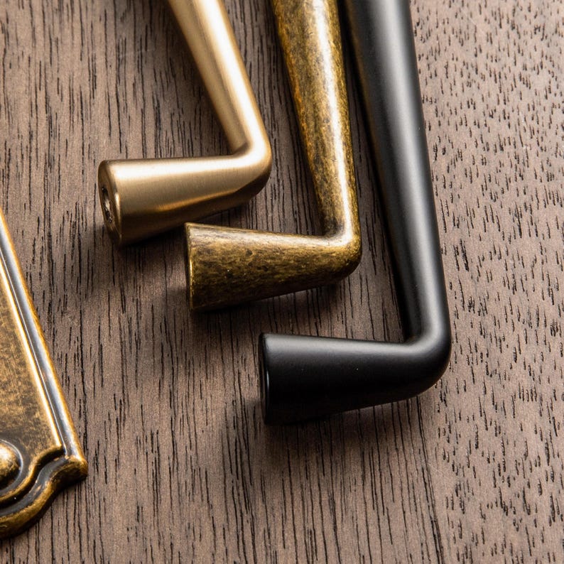 May include: Close-up of three cabinet pulls in different finishes: brass, antique brass, and black. The pulls are L-shaped with a cylindrical grip and are set against a dark wood grain background. The brass pull is a bright gold color.
