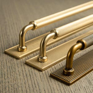 Op de afbeelding: Drie messing kastgrepen met een getextureerd ruitpatroon. Elke greep heeft een gebogen handvat en een rechthoekige achterplaat. De grepen zijn in verschillende tinten goud en brons.