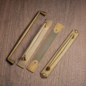 May include: Four antique brass cabinet pulls and handles in various designs. The hardware includes a long, straight handle, and three rectangular pulls with different decorative patterns. The items are arranged on a dark wood surface.