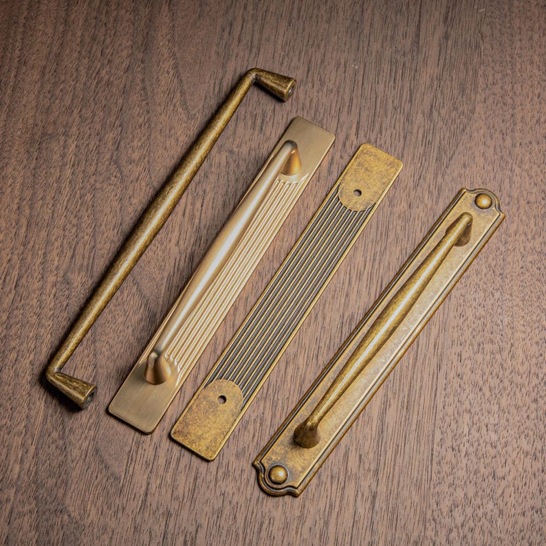 May include: Four antique brass cabinet pulls and handles in various designs. The hardware includes a long, straight handle, and three rectangular pulls with different decorative patterns. The items are arranged on a dark wood surface.