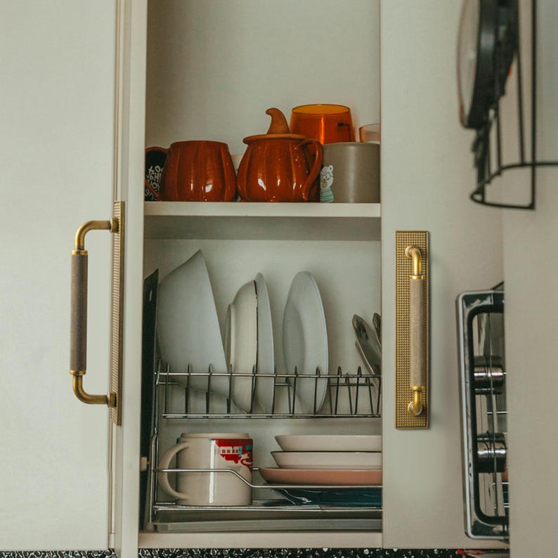 Op de afbeelding: Een witte keukenkast met diverse serviezen en keukengerei. De kastdeuren hebben goudkleurige handgrepen met een bruine grip. Binnenin staan witte borden, oranje mokken en een rood-witte mok.