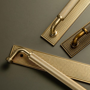 Op de afbeelding: Close-up van drie messing deurgrepen. Elke handgreep heeft een getextureerde, diamantvormige grip en een rechthoekige achterplaat met een bijpassend patroon. De handgrepen zijn gebogen en gemonteerd op een donkergrijze ondergrond.