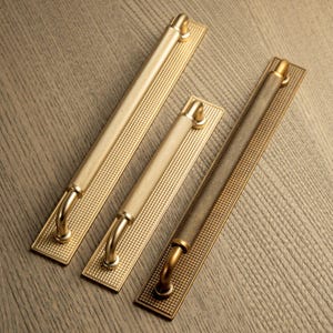 May include: Three gold-toned cabinet pulls with a textured, dotted pattern. Each pull has a rectangular backplate and a curved handle. The pulls vary in size, with the largest measuring approximately 30 cm in length.