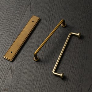 May include: Three brass cabinet hardware pieces on a dark wood surface. The pieces include a rectangular backplate with two holes and two different handle styles: a straight bar and a right-angled bar.