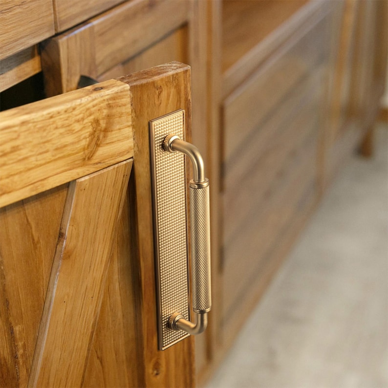 Op de afbeelding: Close-up van een houten kastdeur met een goudkleurige handgreep. De handgreep heeft een rechthoekige achterplaat met een textuurpatroon en een gebogen, cilindrische greep. De kast is gemaakt van lichtgekleurd hout.