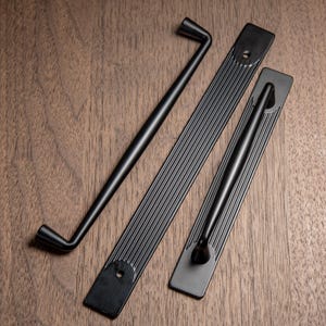 May include: Three black cabinet hardware pieces on a wooden surface. The hardware includes two rectangular pulls with vertical grooves and a curved handle. The hardware is made of metal and has a matte finish.