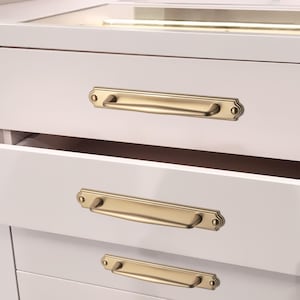 May include: Close-up of a white cabinet with three drawers, each featuring a gold-colored, rectangular handle with decorative end details. The handles are mounted horizontally on the drawer fronts. The top drawer is open, revealing the interior.
