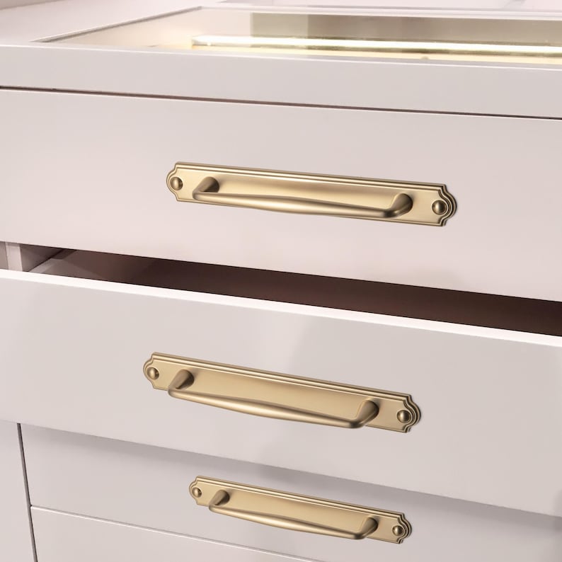May include: Close-up of a white cabinet with three drawers, each featuring a gold-colored, rectangular handle with decorative end details. The handles are mounted horizontally on the drawer fronts. The top drawer is open, revealing the interior.