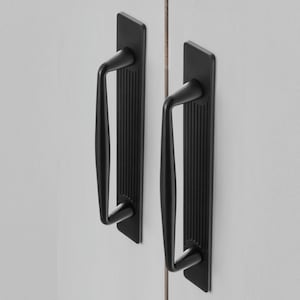 May include: Two black cabinet pulls with a rectangular backplate and a curved handle. The backplate has vertical grooves. The handles are mounted on a white surface.